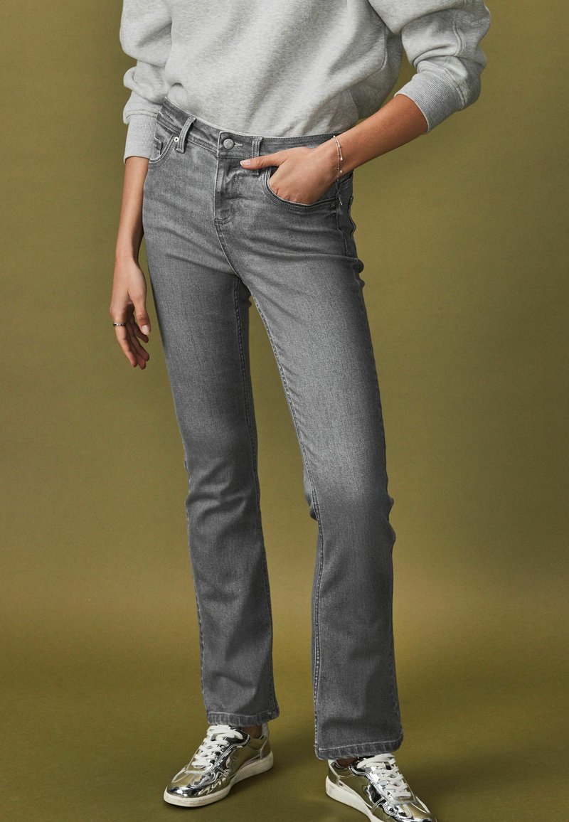 Persona vistiendo jeans ajustados grises, sudadera gris claro, pulsera plateada y zapatillas metálicas plateadas, de pie contra un fondo oliva.