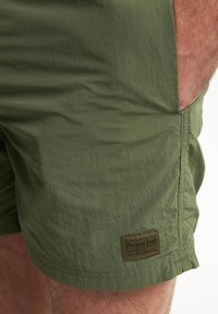 Pantaloni corti verde oliva realizzati in tessuto leggero, con superficie testurizzata e tasche laterali. Include un logo con la scritta "Petrol Ind.".