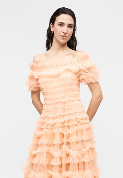 Tumejuustega naine, kes kannab virsikuvärvi õlavaba kleiti, millel on kihilised volangid ja lühikesed varrukad, seisab ühetaustalise heleda tausta ees.