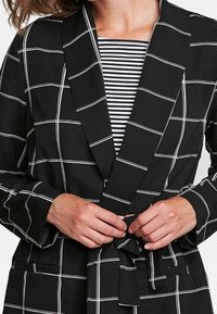 Svart och vit rutigt blazer med slagkrage, tygbälte och långa ärmar, som bärs över en svart och vit randig topp.