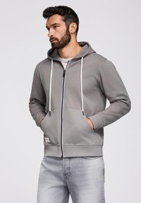 Szara bluza z kapturem na zamek błyskawiczny, z troczkami, przednimi kieszeniami i białym zamkiem. Posiada miękką teksturę i ściągacze przy rękawach. Noszona na jasnej koszuli.