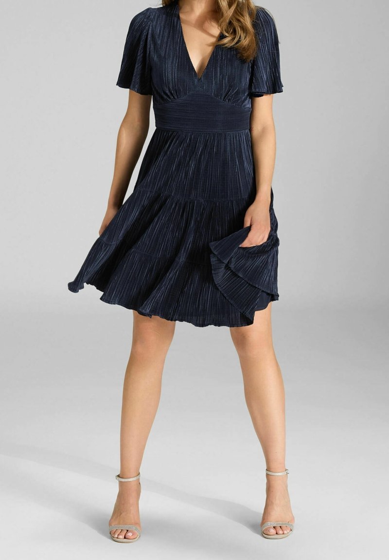 Femme portant une robe plissée bleu marine à manches courtes et col en V, tenant la jupe, associée à des talons beiges à bride à la cheville.