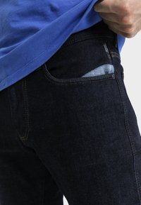 Jeans de mezclilla ajustados de color azul oscuro con forro de bolsillo azul claro, que presentan costuras naranjas y un bolsillo contrastante en la parte frontal.