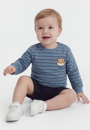 Tout-petit aux cheveux châtain clair portant un pull rayé bleu avec un écusson renard, un short bleu marine et des baskets blanches, assis et souriant.