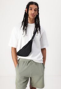 En svart tygslingväska som bärs över en vit t-shirt och ljusgröna shorts. Väskan har ett dragkedjestängning och justerbar rem.