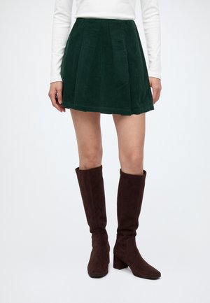 Mujer con minifalda de pana verde oscura, camiseta blanca de manga larga y botas de ante marrón hasta la rodilla, de pie sobre un fondo liso.