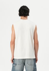 Αμάνικο λευκό βαμβακερό μπλουζάκι με στρογγυλή λαιμόκοψη και άνετη εφαρμογή, διαθέτει καθαρό στρίφωμα και μαλακή υφή, συνδυασμένο με μπλε τζιν σορτς.