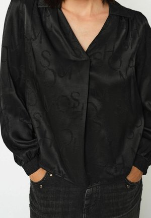 Vrouw draagt een zwarte satijnen blouse met subtiel getextureerde letters en elastische manchetten, handen in de zakken van donkere denim jeans.