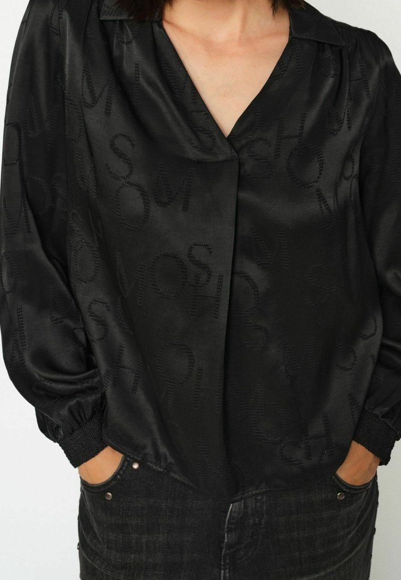 Femme portant un chemisier en satin noir avec des lettres texturées subtiles et des poignets élastiques, mains dans les poches d’un jean en denim foncé.