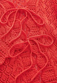 Tissu tricoté rouge avec des motifs en dentelle ouverts, doté de liens distinctifs au centre. Texture douce avec des détails de points complexes.