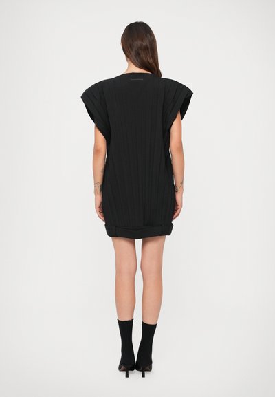 Mulher de costas, vestindo um vestido preto curto com ombreiras, meias pretas até o tornozelo e sapatos altos pretos, em um fundo branco liso.