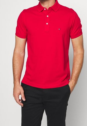 Homme portant un polo à manches courtes rouge vif avec trois boutons et un petit logo sur la poitrine, assorti à un pantalon noir.