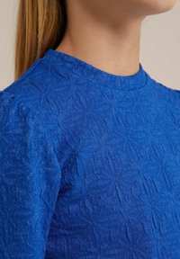 Blå texturerad plagg med rundad halsringning. Tyget visar ett veckat mönster med en subtil glans som reflekterar ljus.