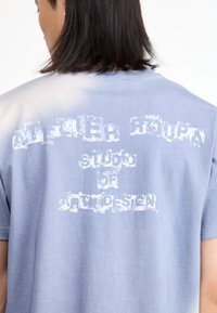 Mėlyna medvilninė marškinė su minkšta tekstūra, ant nugaros yra balti nusidėvėję užrašai "ATELIER ROUPA studija menų dizaino."
