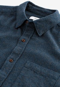 Cămașă din flanel în carouri albastre și maro, cu guler tip button-down, având două butoane maro și un buzunar pe piept. Fabrică moale, texturată.