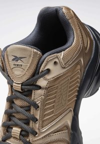 Chaussure de sport marron et noire avec des panneaux texturés, des lacets noirs, un col matelassé et une étiquette "Premier" sur la languette.