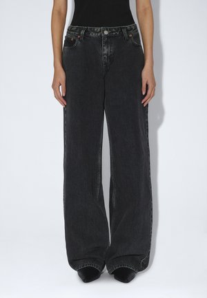 Person iført sorte vidde jeans og sorte spidse sko, stående med hænder afslappet ned langs siderne mod en ensfarvet baggrund.