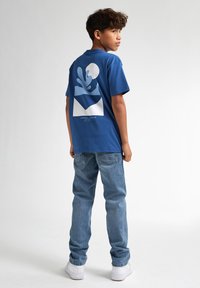 Camiseta de algodón azul con un diseño gráfico que presenta formas abstractas en azul claro y blanco. Combinada con unos vaqueros de mezclilla azul claro y zapatillas blancas.