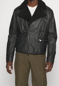 Svart läderjacka med en texturerad yta, stor svart shearlingkrage, flera dragkedjefickor och en figursydd design.