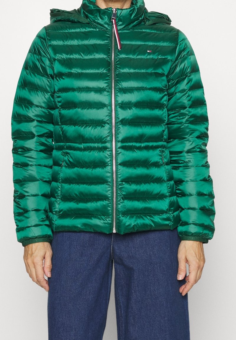 Hombre con una chaqueta acolchada brillante verde con cremallera frontal, capucha y pantalones azul oscuro, de pie contra un fondo liso.