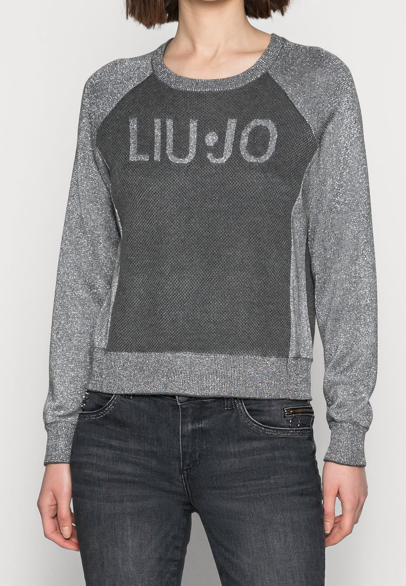 Femme portant un pull gris pailleté à manches longues avec le texte "LIU·JO" sur la poitrine et un jean gris foncé avec une poche zippée.