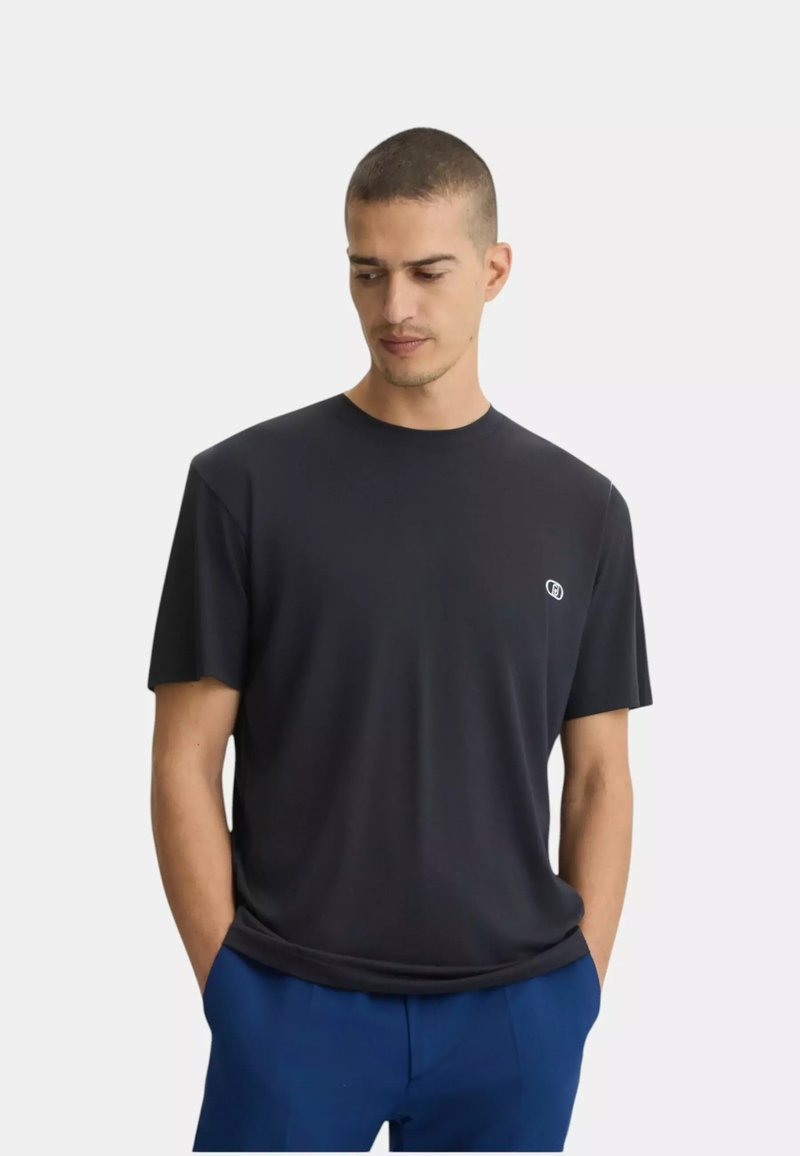 Homme aux cheveux courts portant un t-shirt noir uni et un pantalon bleu, debout les mains dans les poches, regardant vers le bas et sur le côté.