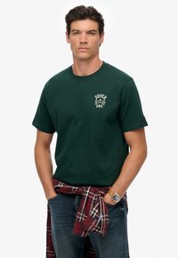 Groen katoenen T-shirt met korte mouwen, voorzien van een wit logo en de tekst 1954. Gemaakt met een rode en geruite flanellen stof om de taille geknoopt.