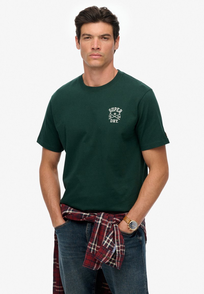 Groen katoenen T-shirt met korte mouwen, voorzien van een wit logo en de tekst 1954. Gemaakt met een rode en geruite flanellen stof om de taille geknoopt.