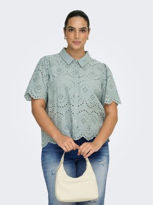 Femme portant une chemise bleu clair à œillets et boutons, et un jean bleu déchiré, tenant un petit sac à main beige devant un fond uni.