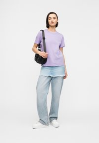 Persoon die een lavendel t-shirt draagt, wijde lichtblauwe spijkerbroek met een versleten denim rok eroverheen, witte sneakers en een zwarte schoudertas.