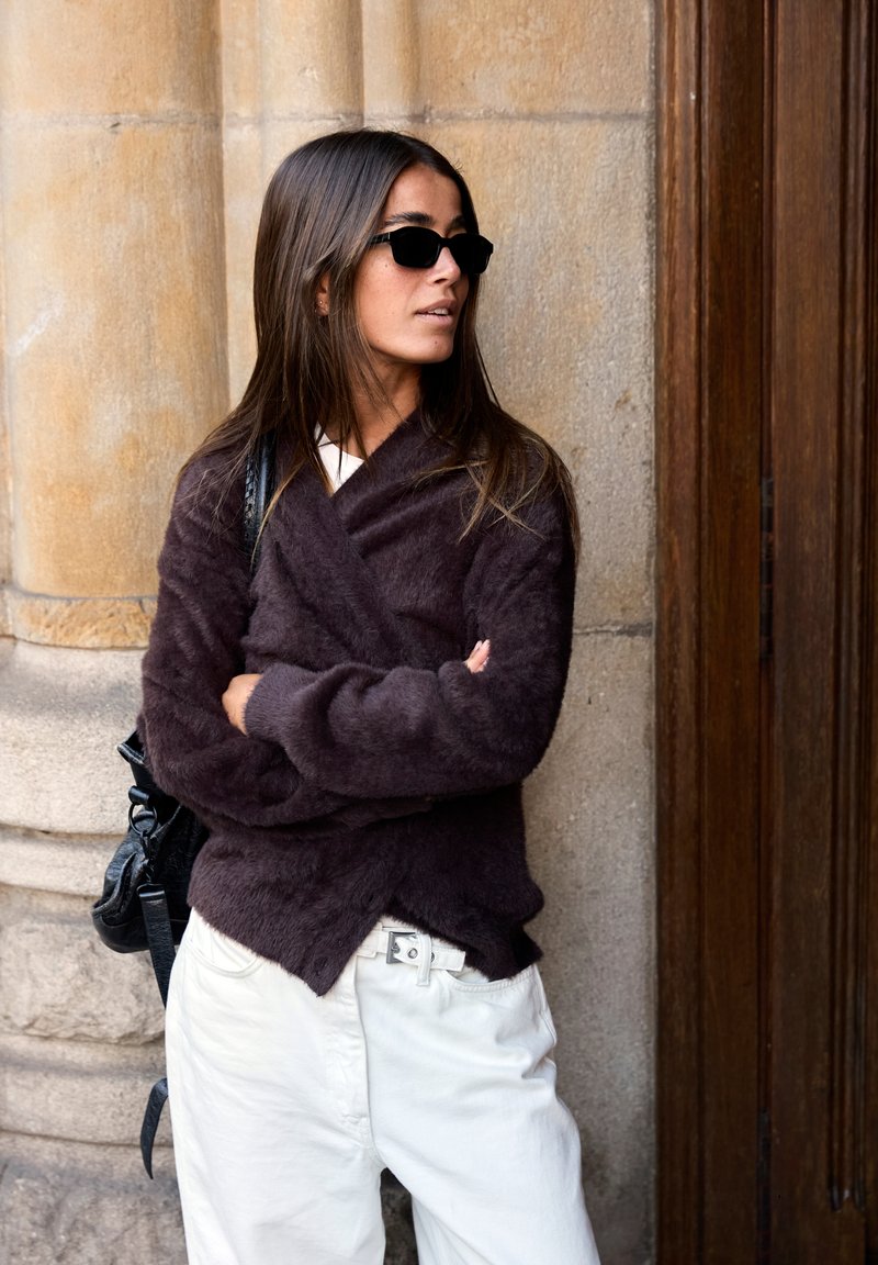 Mörkbrun, fluffig omslagsjumper, vita byxor, svart handväska och svarta solglasögon. Slät yta med avslappnad passform och asymmetrisk fåll.