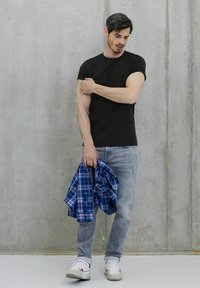 Černé přiléhavé tričko, světle šedé džíny, drží modrou kostkovanou košili. Tenisky s bílými a vínovými akcenty. Betonové pozadí.