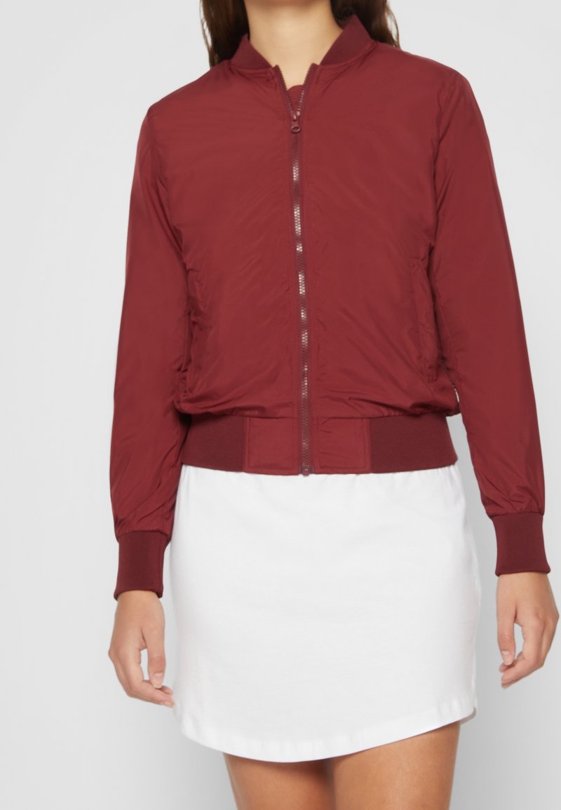 Femme portant une veste bomber bordeaux zippée avec des poignets et une taille côtelés, sur une jupe blanche, debout devant un fond uni.
