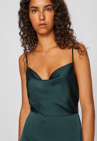 Kvinne med krøllete mørkt hår iført en mørkegrønn sateng ermeløs kjole med tynne stropper, mot en ensfarget lys bakgrunn.