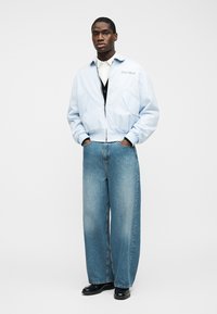 Homme debout, les mains dans les poches, portant une veste bleu clair, une chemise blanche, un gilet noir, un jean bleu à jambes larges et des chaussures noires sur un fond blanc.