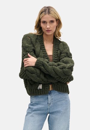 Duży, ciemnozielony sweter kardigan z grubymi warkoczami, głębokim dekoltem w szpic i prążkowanym dołem. Noszony na jasnej bluzce i zestawiony z niebieskimi dżinsami.