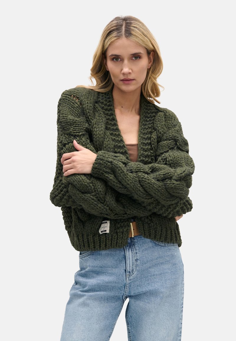 Duży, ciemnozielony sweter kardigan z grubymi warkoczami, głębokim dekoltem w szpic i prążkowanym dołem. Noszony na jasnej bluzce i zestawiony z niebieskimi dżinsami.