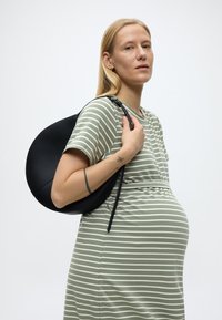 Έγκυος γυναίκα με φόρεμα με πράσινες και λευκές ρίγες, κρατώντας μια μαύρη στρογγυλή τσάντα στον ώμο της, μπροστά σε απλό φόντο.