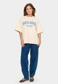 Béžové oversized tričko s modrým nápisem "SANTA MONICA STATE BEACH," spárované s tmavě modrými džíny a plochými botami s leopardím vzorem.