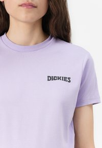 Kratka majica s kratkimi rokavi svetlo vijolične barve, izdelana iz bombaža, z črnim napisom "DICKIES" na levi prsni strani.