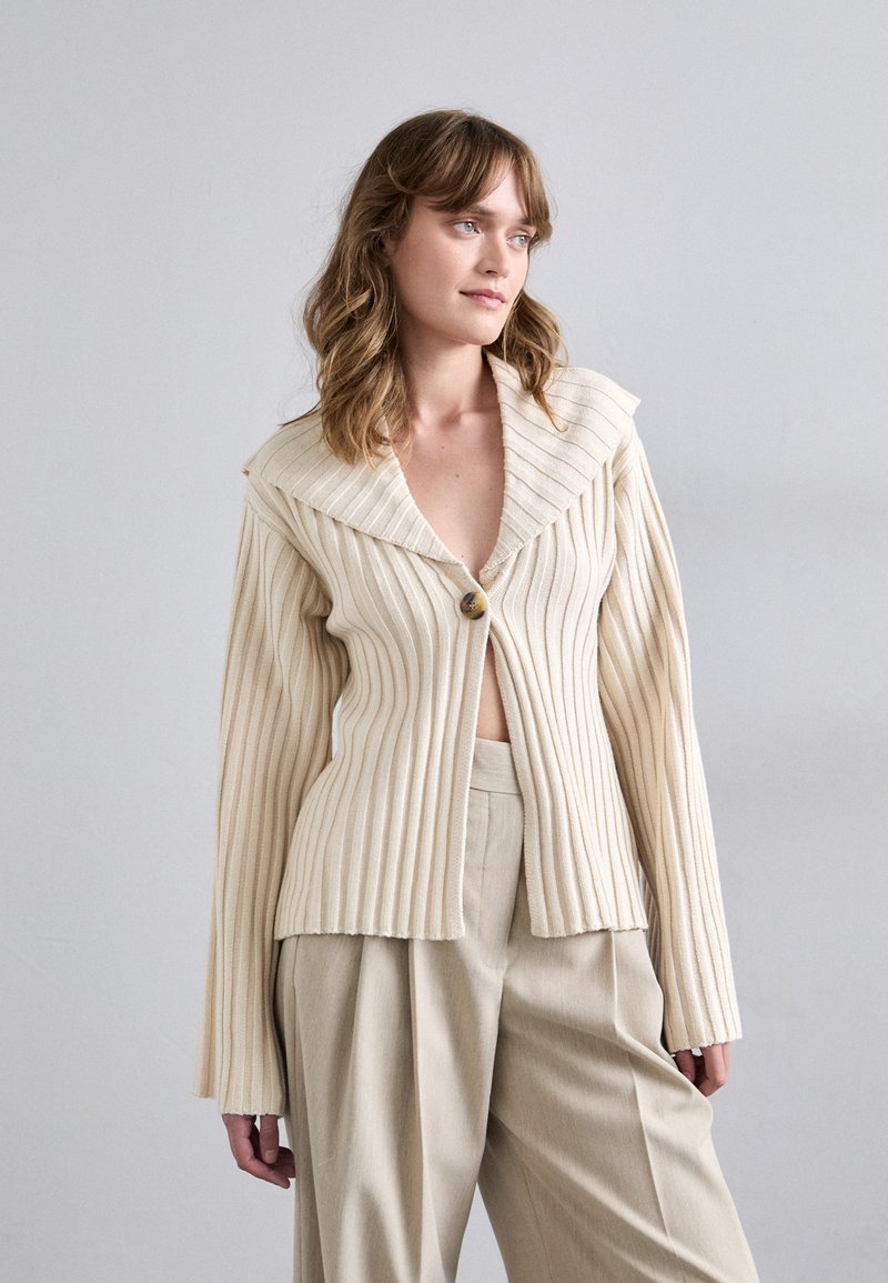 Cream ribbed cardigan with a wide collar and single button closure. Long sleeves and textured fabric, styled open over beige trousers.