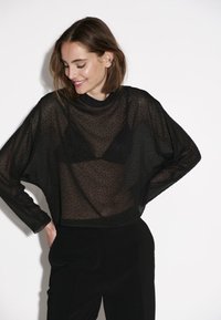 Schwarzes, transparentes Oberteil mit gepunktetem Muster, lockerer Passform, rundem Ausschnitt und langen Ärmeln. Tragen über einem schwarzen Bralette und kombiniert mit schwarzen Hosen.