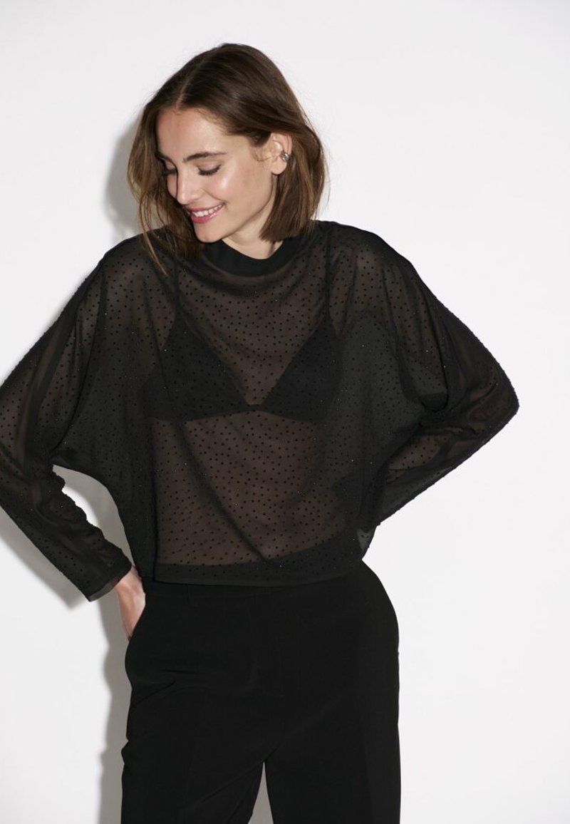 Schwarzes, transparentes Oberteil mit gepunktetem Muster, lockerer Passform, rundem Ausschnitt und langen Ärmeln. Tragen über einem schwarzen Bralette und kombiniert mit schwarzen Hosen.