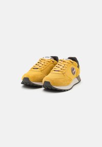 Sneakers din suede galben și material textil, cu accente negre, detaliu cu logo rotund, talpă texturată și design cu șireturi. Profil subțire cu o talpă intermediară contrastantă.