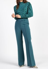Donna che indossa pantaloni a vita alta e gamba larga di colore teal, una maglietta teal con motivo floreale e maniche arricciate, e scarpe metalliche a punta, in piedi.