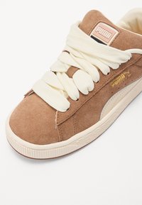 Brown suede sneaker with cream-colored laces, a textured rubber sole, and a gold-printed logo on the side and tongue.
