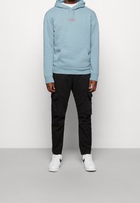 Person som bär en ljusblå hoodie, svarta cargobyxor och vita Nike-sneakers, stående mot en enfärgad ljusgrå bakgrund.