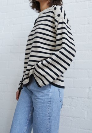 Femme portant un pull rayé noir et blanc avec des boutons sur l'épaule, associé à un jean bleu clair, debout contre un mur en briques blanches.