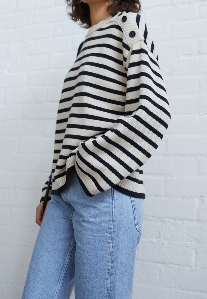 Femme portant un pull rayé noir et blanc avec des boutons sur l'épaule, associé à un jean bleu clair, debout contre un mur en briques blanches.