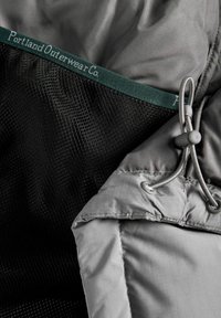 Szara tkanina wierzchnia o gładkiej fakturze, z siateczkowym wykończeniem i ciemnozieloną metką, na której widnieje napis "Portland Outerwear Co." oraz detale ze sznureczkiem.